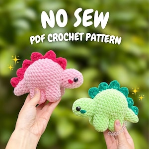 Puede incluir: Dos juguetes de dinosaurio de peluche hechos de hilo suave, uno en rosa claro con púas rosa oscuro y otro en verde claro con púas verde oscuro. El texto NO SEW PDF CROCHET PATTERN aparece sobre los juguetes, que tienen ojos negros.