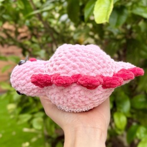 May include: A small, soft, light pink crocheted plush toy shaped like a dinosaur. It features a row of dark pink, rounded spikes along its back and small black eyes. The text @AQcrochets is visible on the side of the plush body.