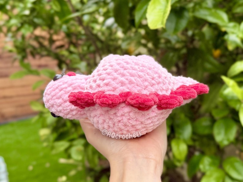 May include: A small, soft, light pink crocheted plush toy shaped like a dinosaur. It features a row of dark pink, rounded spikes along its back and small black eyes. The text @AQcrochets is visible on the side of the plush body.