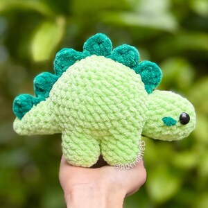 May include: A light green crocheted dinosaur plush toy with a textured yarn finish. The dinosaur features a row of dark green, rounded spikes along its back, a small dark green patch on its cheek, and a single black plastic eye on its side.