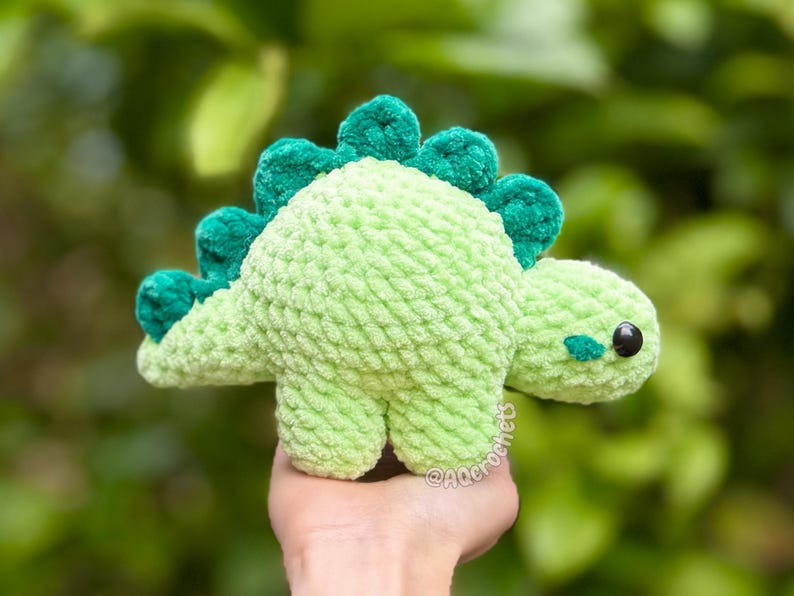 May include: A light green crocheted dinosaur plush toy with a textured yarn finish. The dinosaur features a row of dark green, rounded spikes along its back, a small dark green patch on its cheek, and a single black plastic eye on its side.