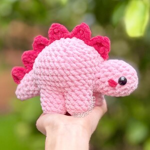 May include: A light pink crocheted dinosaur plush toy with a row of dark pink spikes along its back. The dinosaur features a rounded body, four short legs, and a small head with a black plastic eye and a small pink stitched cheek detail.