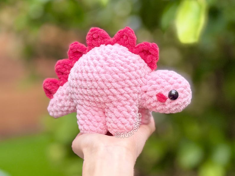 May include: A light pink crocheted dinosaur plush toy with a row of dark pink spikes along its back. The dinosaur features a rounded body, four short legs, and a small head with a black plastic eye and a small pink stitched cheek detail.