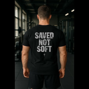 May include: Black t-shirt with the text "SAVED NOT SOFT" in distressed white lettering and a small cross graphic. The shirt is short-sleeved and appears to be made of a soft material. The person is wearing black shorts.