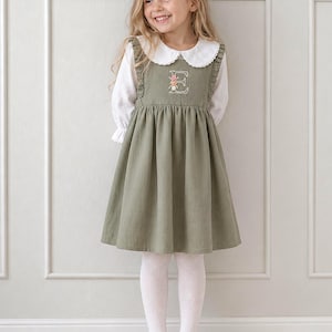 Könnte beinhalten: Ein salbeigrünes Trägerkleid mit gerüschten Schulterträgern und einem aufgestickten Buchstaben E mit Blumenakzenten auf der Brust. Das Kleid wird über einer weißen langärmeligen Bluse mit rundem Kragen getragen, kombiniert mit weißen Strumpfhosen und braunen Schuhen.
