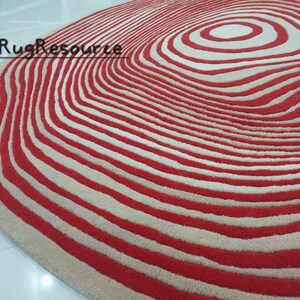 May include: A red and cream colored rug with a circular pattern of concentric circles. The rug has a wavy edge.