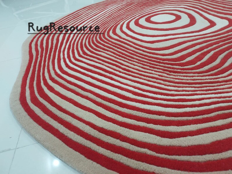 May include: A red and cream colored rug with a circular pattern of concentric circles. The rug has a wavy edge.