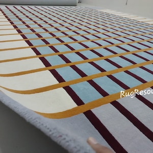 Puede incluir: Primer plano de una alfombra rayada blanca, azul y burdeos con una raya amarilla que corre en diagonal a través de la alfombra. La alfombra está enrollada y el borde de la alfombra es visible. El texto "RugResource" es visible en la esquina inferior derecha de la imagen.