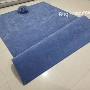 May include: A blue rectangular rug with a rolled-up section. A small, blue, decorative item sits on the rug. The rug is made of a soft material and is displayed on a tiled floor.