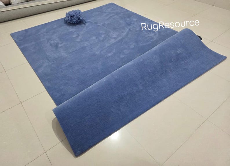May include: A blue rectangular rug with a rolled-up section. A small, blue, decorative item sits on the rug. The rug is made of a soft material and is displayed on a tiled floor.