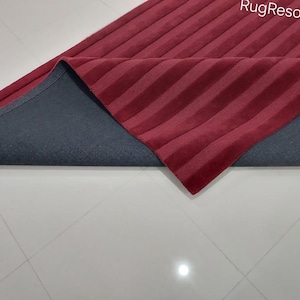 Op de afbeelding: Een rood en grijs gestreept tapijt met een zachte, pluche textuur. Het tapijt is dubbelgevouwen, waardoor de grijze achterkant zichtbaar wordt.