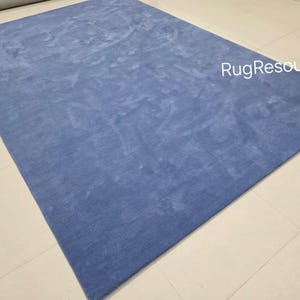 May include: A large, rectangular, solid blue rug. The rug has a soft, textured appearance and is placed on a light-colored tiled floor. The text "RugResource" is visible in the upper right corner.