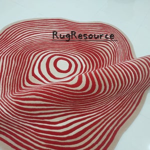 May include: A red and cream colored rug with a circular pattern of concentric circles. The rug is made of wool and has a soft, plush texture.