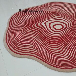 May include: A red and cream colored round rug with a wavy, organic shape and a concentric circle pattern. The rug has a modern, abstract design.