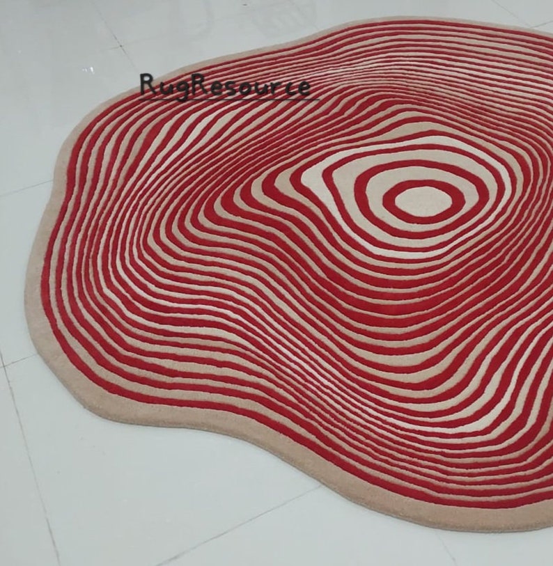 May include: A red and cream colored round rug with a wavy, organic shape and a concentric circle pattern. The rug has a modern, abstract design.