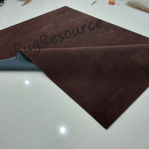 K&ouml;nnte beinhalten: Ein rechteckiger, dunkelbrauner Teppich mit einer gefalteten Ecke, die eine blaue Unterseite zeigt. Der Teppich liegt auf einem wei&szlig;en Fliesenboden. Der Text "Rug Resource" ist teilweise auf der Oberfl&auml;che des Teppichs sichtbar.