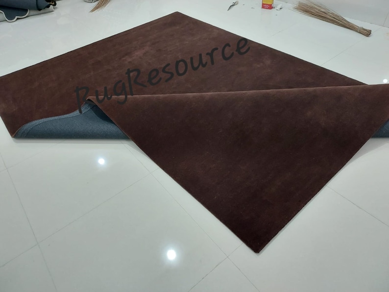 K&ouml;nnte beinhalten: Ein rechteckiger, dunkelbrauner Teppich mit einer gefalteten Ecke, die eine blaue Unterseite zeigt. Der Teppich liegt auf einem wei&szlig;en Fliesenboden. Der Text "Rug Resource" ist teilweise auf der Oberfl&auml;che des Teppichs sichtbar.