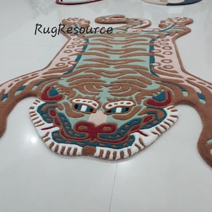 Op de afbeelding: Een decoratief tapijt in de vorm van een tijger, met een gedetailleerd ontwerp. Het tapijt heeft een bruin, beige en blauwgroen kleurenschema met witte en rode accenten. Het tapijt ligt op een witte ondergrond.