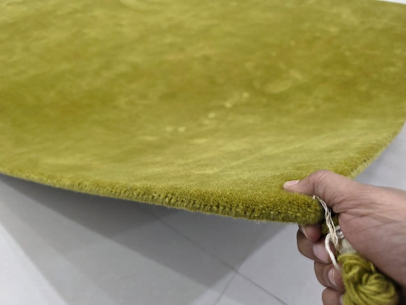 K&ouml;nnte beinhalten: Nahaufnahme eines weichen, olivgr&uuml;nen Teppichs. Der Teppich hat eine weiche Textur und eine leicht gebogene Kante. Eine Hand hebt den Teppich an und zeigt die Unterseite und die Konstruktion der Kante. Der Teppich ist ein leuchtendes Gr&uuml;n.