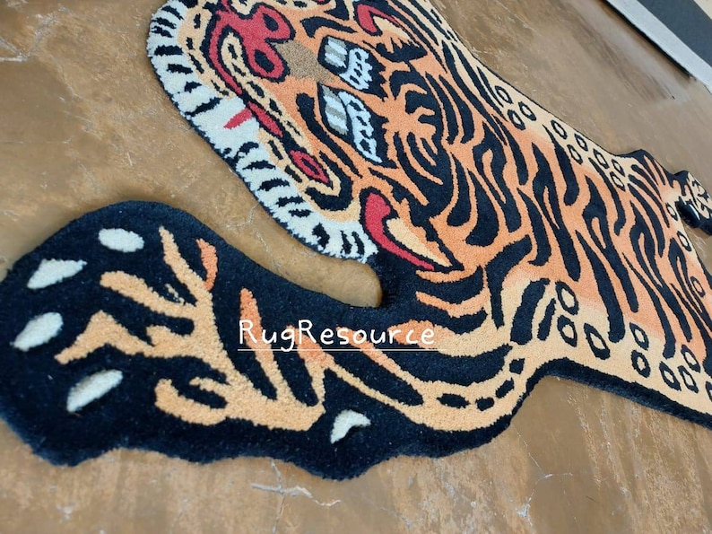 Op de afbeelding: Een tapijt in de vorm van een tijger, met oranje, zwarte en witte kleuren. Het tapijt heeft een gedetailleerd ontwerp met strepen en gezichtskenmerken, waaronder ogen en een neus. De tekst "RugResource" is zichtbaar op het tapijt.