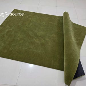 May include: A rectangular olive green rug with a rolled-up edge, revealing a dark backing. The rug is on a white tiled floor. The text "RugResource" is in the upper left corner.