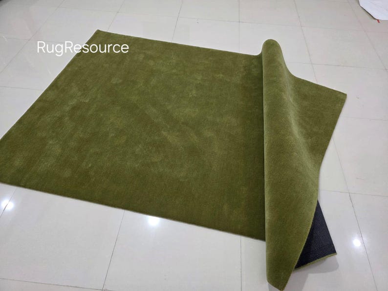 May include: A rectangular olive green rug with a rolled-up edge, revealing a dark backing. The rug is on a white tiled floor. The text "RugResource" is in the upper left corner.