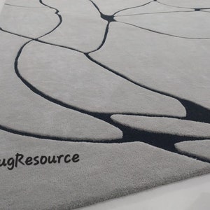 Puede incluir: Primer plano de una alfombra abstracta gris y negra con superficie texturizada. La alfombra tiene un dise&ntilde;o moderno con un patr&oacute;n de l&iacute;neas y formas negras sobre un fondo gris. El texto "RugResource" es visible en la esquina inferior izquierda de la imagen.