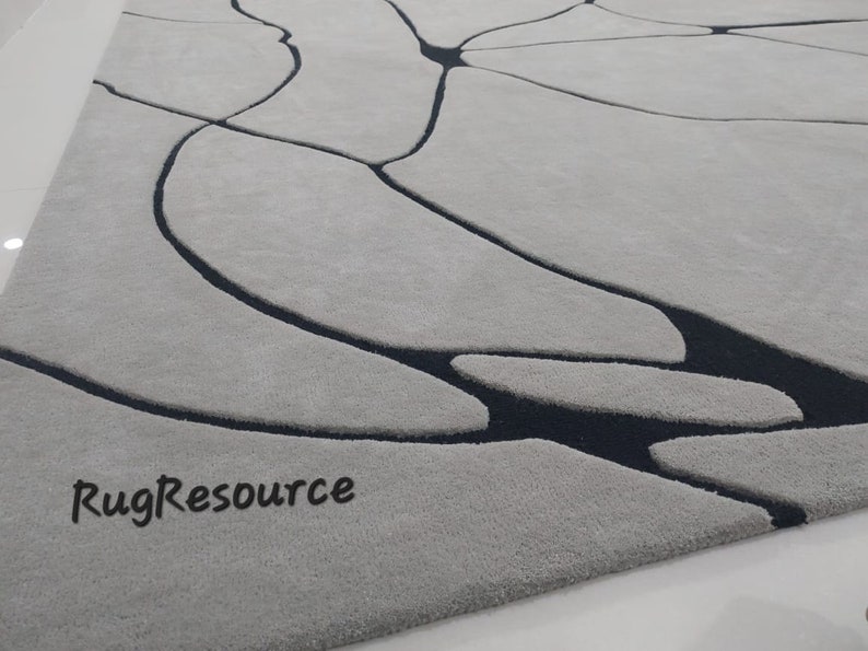 Puede incluir: Primer plano de una alfombra abstracta gris y negra con superficie texturizada. La alfombra tiene un dise&ntilde;o moderno con un patr&oacute;n de l&iacute;neas y formas negras sobre un fondo gris. El texto "RugResource" es visible en la esquina inferior izquierda de la imagen.