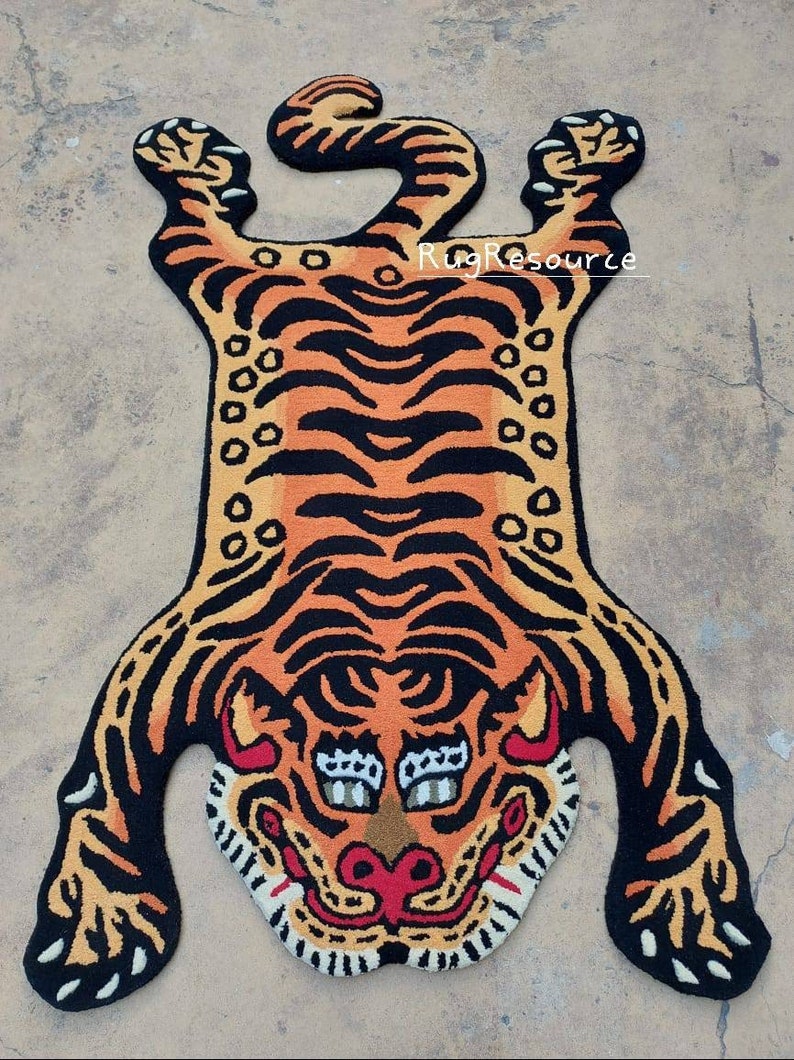 Op de afbeelding: Een tijgervormig tapijt met een gedetailleerd ontwerp. Het tapijt heeft oranje en zwarte strepen, een tijgergezicht met witte tanden en een rode mond. Het tapijt is ontworpen om op een tijgervel te lijken.