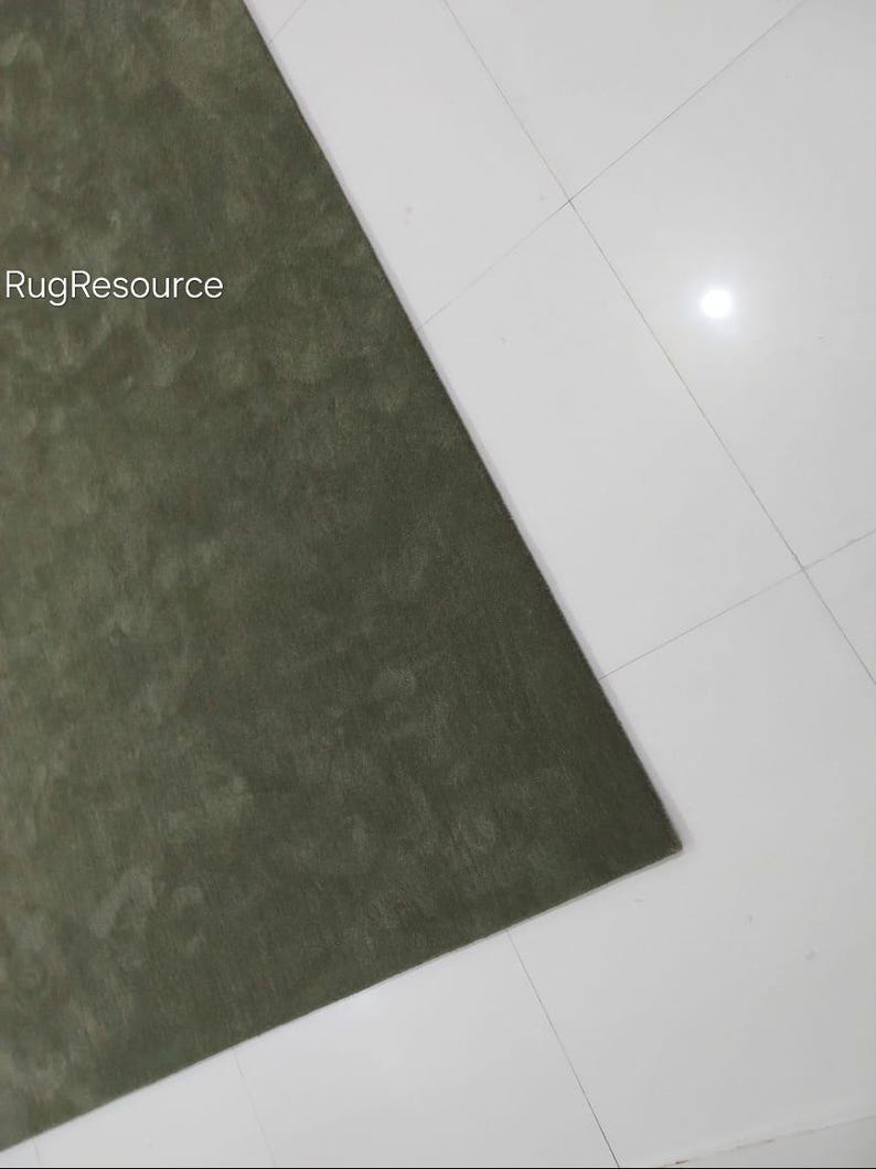 May include: A section of a green rug with a textured surface. The rug is positioned diagonally against a white tiled floor. The text "RugResource" is visible in the upper left corner.