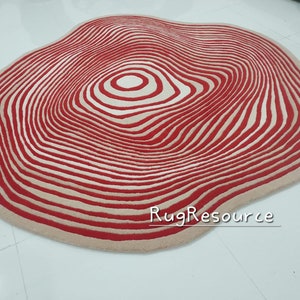 May include: A large, round, cream-colored rug with a red and white concentric circle pattern. The rug has a wavy, organic shape.