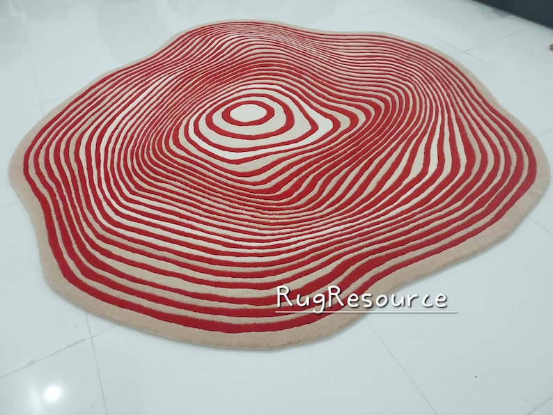 May include: A large, round, cream-colored rug with a red and white concentric circle pattern. The rug has a wavy, organic shape.