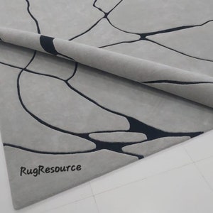 Puede incluir: Primer plano de una alfombra gris con un dise&ntilde;o abstracto negro. La alfombra est&aacute; enrollada y el logotipo de RugResource es visible en la esquina inferior derecha.