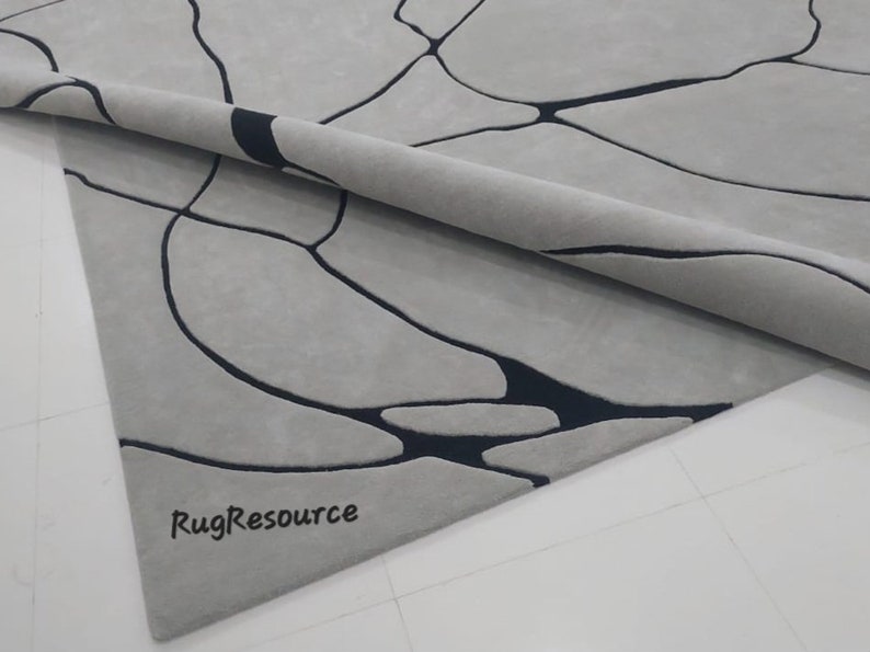 Puede incluir: Primer plano de una alfombra gris con un dise&ntilde;o abstracto negro. La alfombra est&aacute; enrollada y el logotipo de RugResource es visible en la esquina inferior derecha.