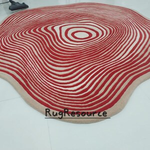 May include: A large, cream-colored rug with a red and white concentric circle pattern. The rug has a wavy, organic shape.