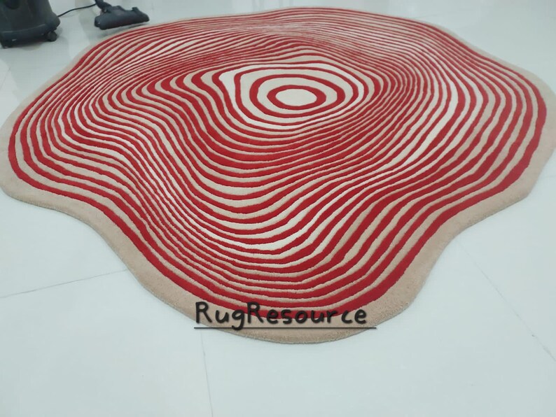 May include: A large, cream-colored rug with a red and white concentric circle pattern. The rug has a wavy, organic shape.
