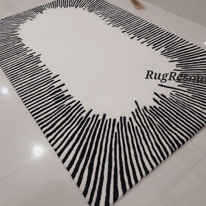 May include: A white rug with a black oval border made of vertical lines. The rug has a modern design.