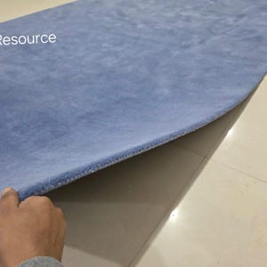 May include: A light blue rectangular rug with a soft, plush texture. The rug is being lifted to reveal its underside. The text "RugResource" is visible in the upper left corner. The rug appears to be made of a soft material.