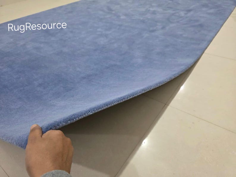 May include: A light blue rectangular rug with a soft, plush texture. The rug is being lifted to reveal its underside. The text "RugResource" is visible in the upper left corner. The rug appears to be made of a soft material.