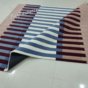 May include: A white, blue, and pink striped rug with a geometric pattern. The rug is folded in half and the underside is visible. The rug is made of a soft, plush material.