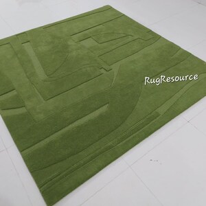 May include: A green area rug with a geometric pattern. The rug has a raised texture and is made of wool. The rug is on a white surface. RugResource is printed on the rug.