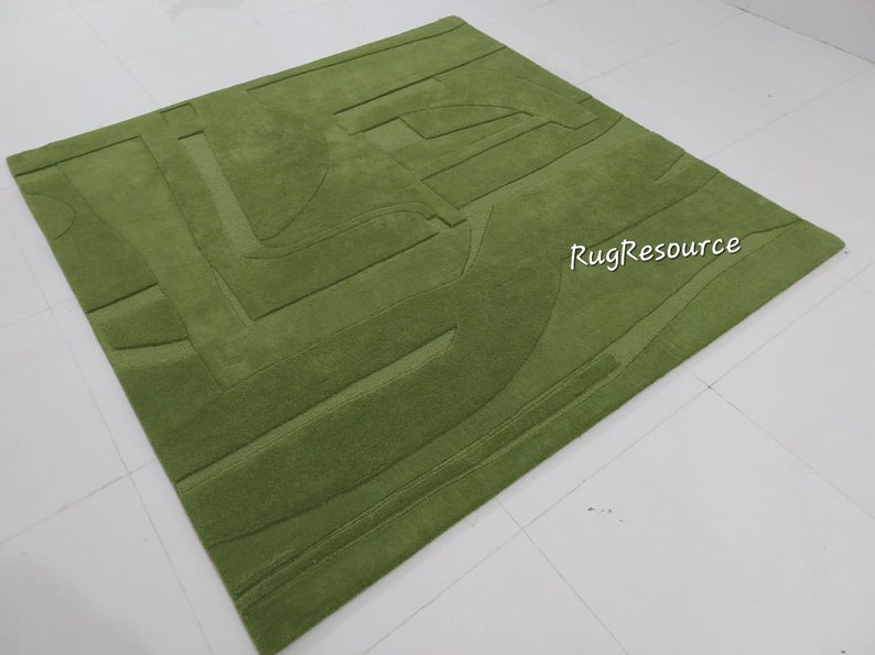 May include: A green area rug with a geometric pattern. The rug has a raised texture and is made of wool. The rug is on a white surface. RugResource is printed on the rug.