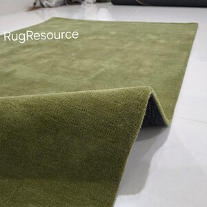 May include: A rectangular olive green rug with a soft, textured surface. The rug is partially rolled up, revealing a dark backing. The text "RugResource" is visible in the upper left corner. The rug is on a white tile floor.