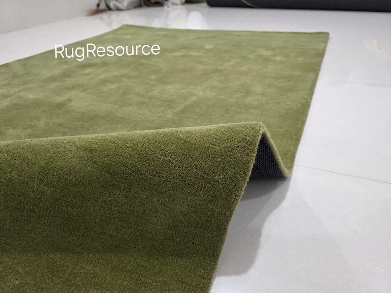 May include: A rectangular olive green rug with a soft, textured surface. The rug is partially rolled up, revealing a dark backing. The text "RugResource" is visible in the upper left corner. The rug is on a white tile floor.