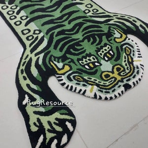 Op de afbeelding: Een groen en zwart tapijt in de vorm van een tijger met een gedetailleerd ontwerp. Het tapijt heeft een tijgergezicht en -lichaam met strepen en vlekken. Het tapijt is gemaakt van een zacht, pluche materiaal en is geschikt voor een woon- of slaapkamer.