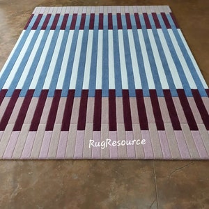 May include: A striped rug with alternating bands of white, light blue, burgundy, and pink. The rug is made of a soft, plush material and has a modern, geometric design. The rug is photographed on a brown floor. The text "RugResource" is visible in the bottom center of the rug.