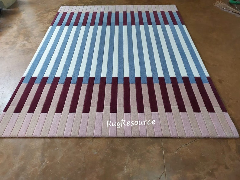 May include: A striped rug with alternating bands of white, light blue, burgundy, and pink. The rug is made of a soft, plush material and has a modern, geometric design. The rug is photographed on a brown floor. The text "RugResource" is visible in the bottom center of the rug.
