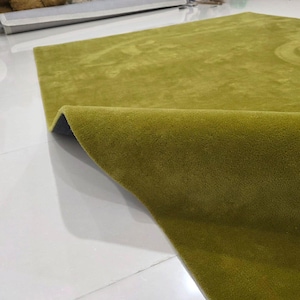 K&ouml;nnte beinhalten: Nahaufnahme eines gr&uuml;nen Teppichs mit gerolltem Rand. Der Teppich hat eine weiche, pl&uuml;schige Textur und eine einfarbige gr&uuml;ne Farbe. Der Teppich liegt auf einer wei&szlig;en Oberfl&auml;che.