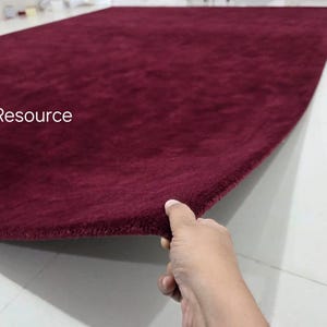 K&ouml;nnte beinhalten: Ein rechteckiger Teppich in tiefem Burgunderrot mit einer weichen, flauschigen Textur. Der Teppich ist an einer Ecke angehoben, um seine Dicke und seinen Flor zu zeigen. Der Text RugResource ist in Wei&szlig; auf der linken Seite des Teppichs gedruckt.