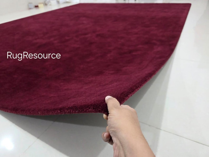 K&ouml;nnte beinhalten: Ein rechteckiger Teppich in tiefem Burgunderrot mit einer weichen, flauschigen Textur. Der Teppich ist an einer Ecke angehoben, um seine Dicke und seinen Flor zu zeigen. Der Text RugResource ist in Wei&szlig; auf der linken Seite des Teppichs gedruckt.